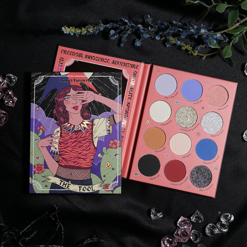 An eye palette with 12 shades including matte, shimmer, and glitter finishes, with a graphic of a woman with a tarot card in the background.