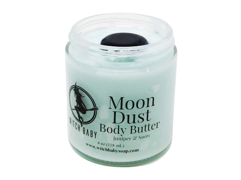 Moon Dust Body Butter is pictured on a white background. It is an icy blue body butter in a glass jar without a lid to show that it is topped with a blue goldstone crystal. Label is clear and reads: Moon Dust Body Butter. Juniper & Snow. 4 oz (118 mL). www.witchbabysoap.com